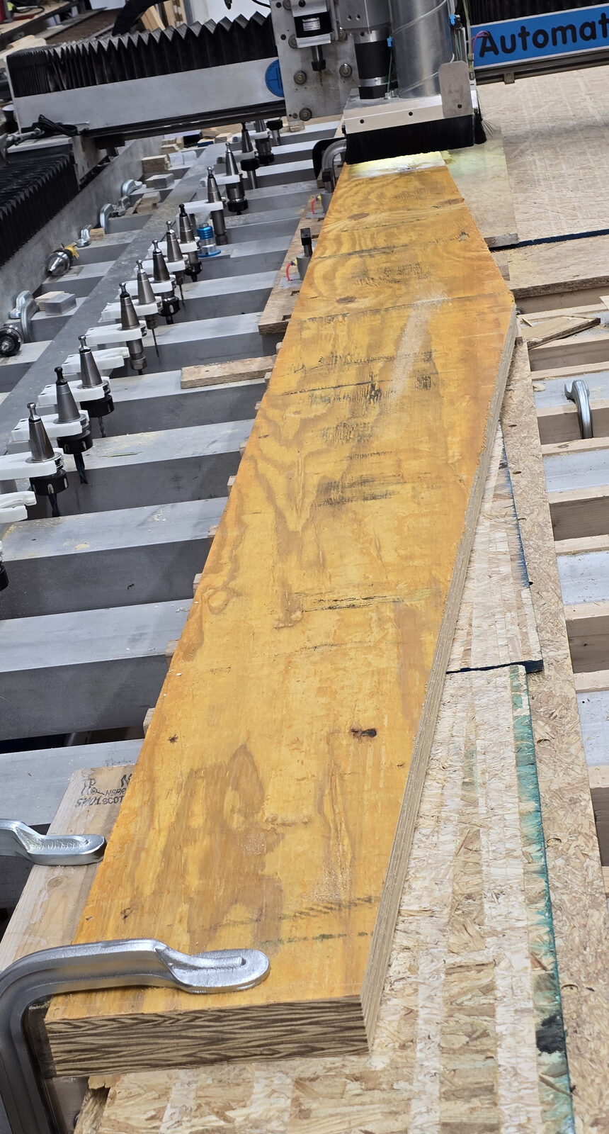 Long LVL plank clamped along the bed of a large CNC router, with the tool changer beside it